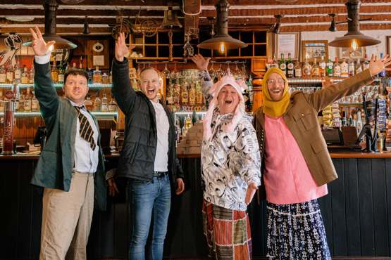 Four men dressed in various outfits stand at a bar with arms outstretched